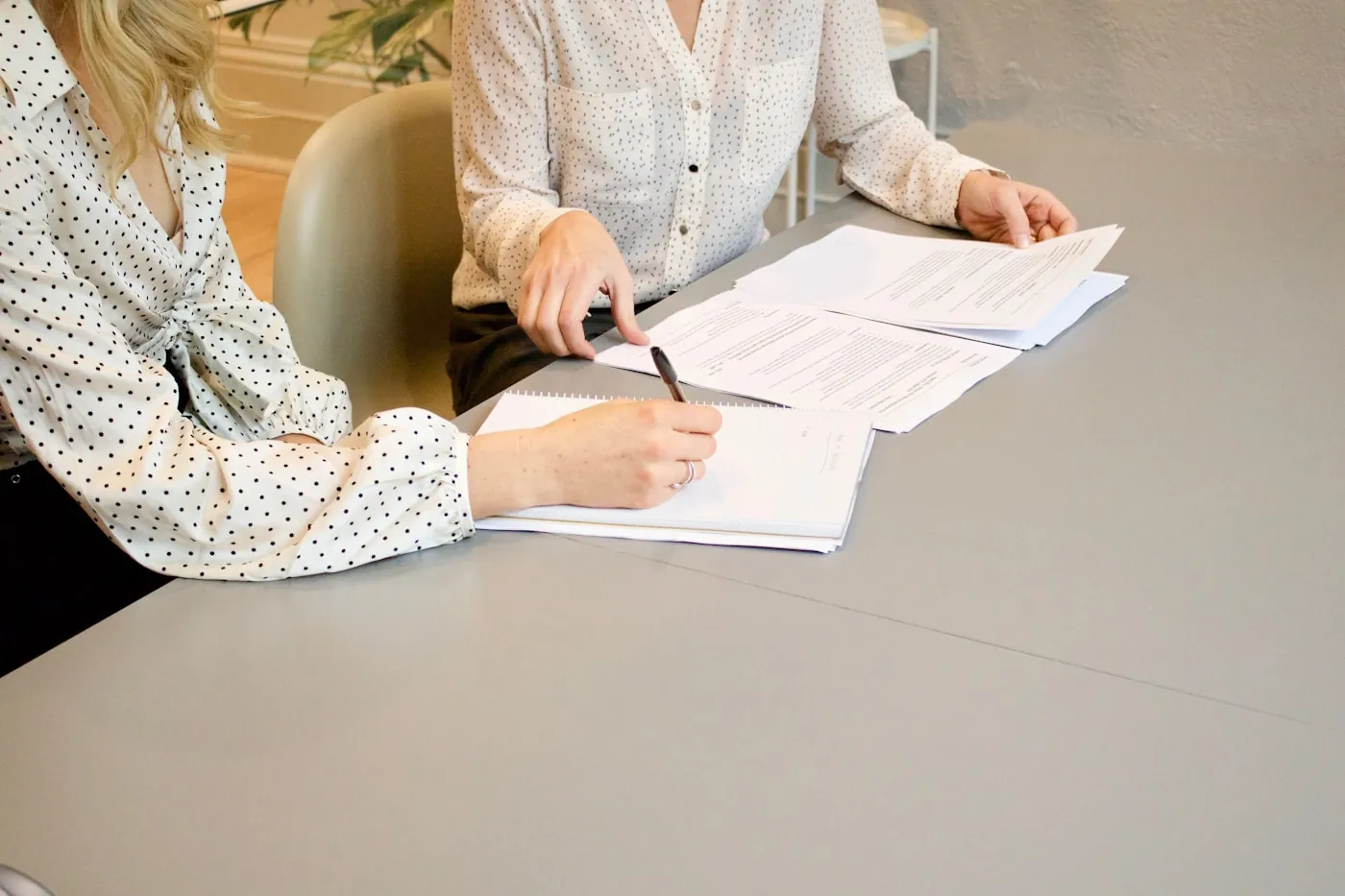 Duas pessoas a uma mesa de escritório, com um contrato impresso aberto entre elas, uma delas anotando num caderno.